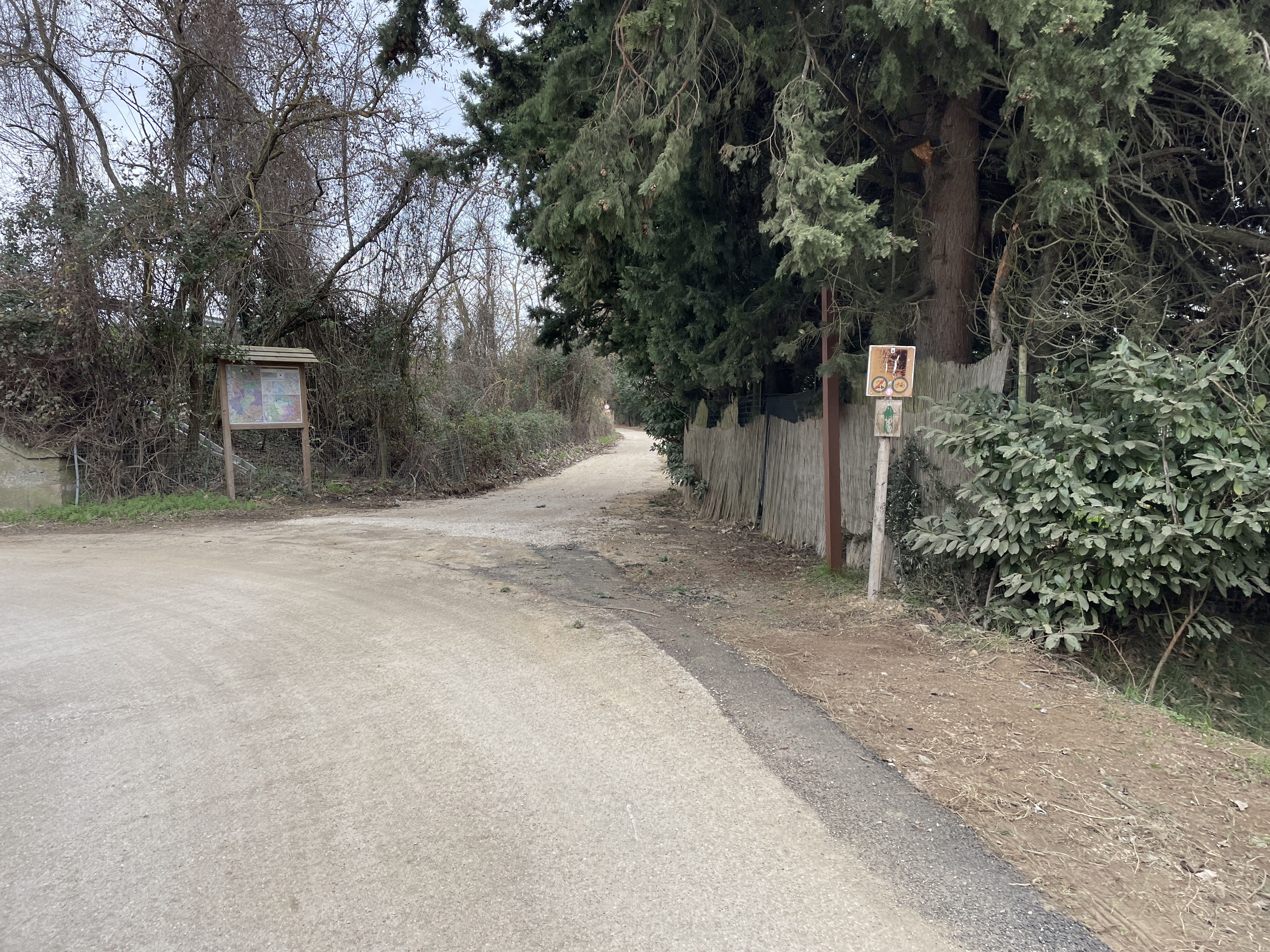 Crossroads between paved road and dirt track. Straw fence on left and map on right side. Road elevated to the path.